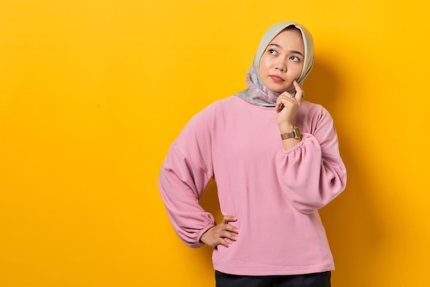 Pensive young Asian woman in pink shirt looks seriously thinking about a question on yellow background