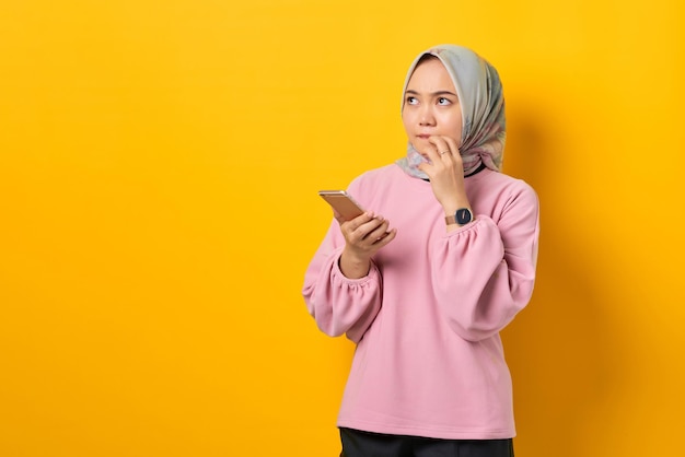 Pensive young Asian woman in pink shirt using smartphone thinking about a question on yellow background