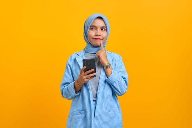 Pensive young Asian woman using mobile phone with fingers on chin over yellow background