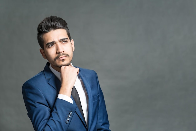 Pensive young attractive man in a blue suit with hand on chin on a gray background