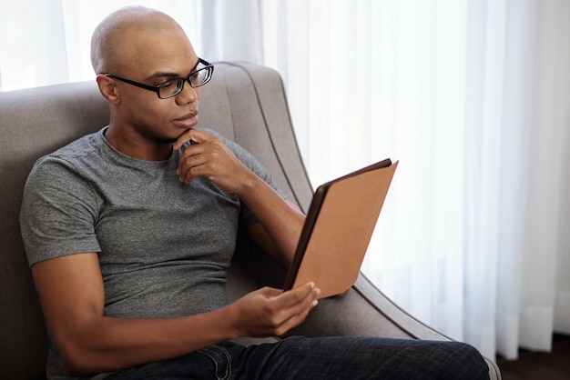 Pensive young bald Black man in glasses sitting in armchair and reading intresting book on tablet computer