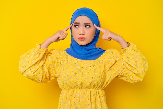 Pensive young beautiful asian muslim woman trying hard to\
remember something isolated over yellow background