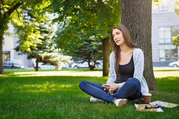 Pensive young brunette asian girl relaxing on a grass and drinking coffee in the park during the day. Lifestyle and technology concept