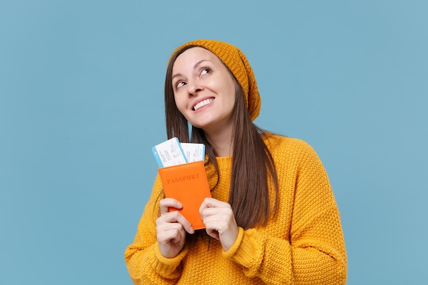 Pensive young brunette woman girl in sweater and hat posing isolated on blue background studio portrait. People lifestyle concept. Mock up copy space. Hold passport boarding pass tickets looking up.