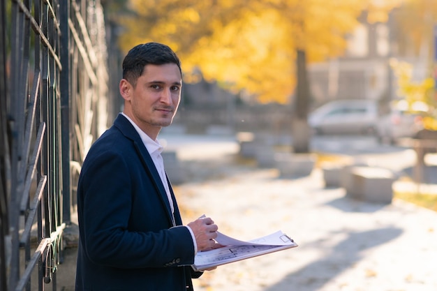 Pensive young businessman man in classic tie suit standing outside hold clipboard with papers read document.