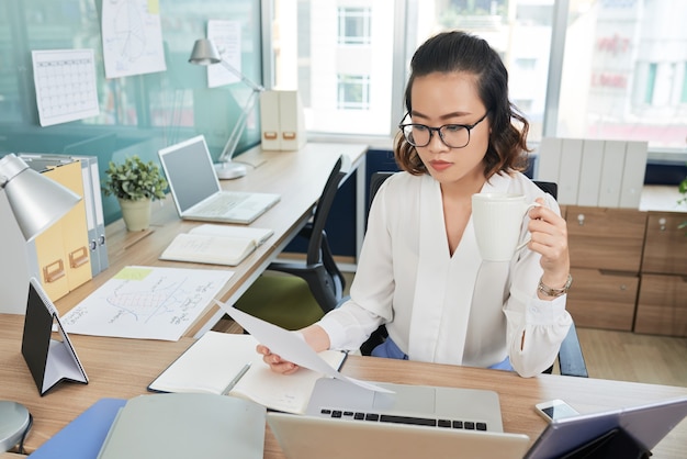 Pensive young businesswoman drinking coffee and reading contract or report when working in modern office