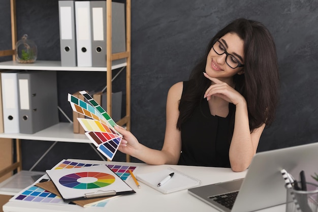 Pensive young female designer in casual working with color palette in modern workplace interior, copy space