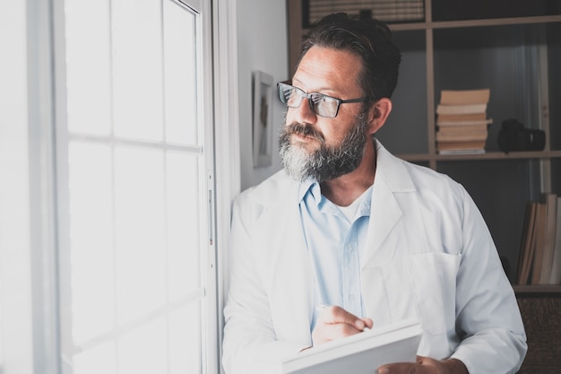 Pensive young male caucasian doctor in white medical uniform look in window distance thinking or pondering, serious man GP plan future career or success in medicine, visualize at workplace writing