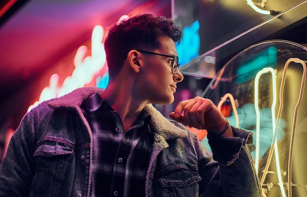 A pensive young man fashionably dressed leaning on an illuminated signboard in the street at night.