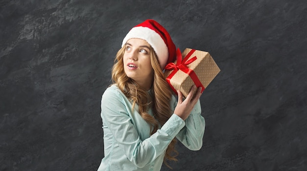 Pensive young man in santa hat shaking gift box and thinking about what is inside, Christmas party and surprise concept, black studio background