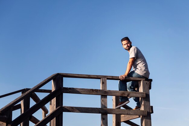 Pensive young man sitting on a wooden railing with blue sky in the background concept of leisure and relax copy space for text