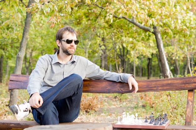 Pensive young man in sunglasses sitting on a bench in woodland with a chessboard alongside him staring off into the distance deep in thought