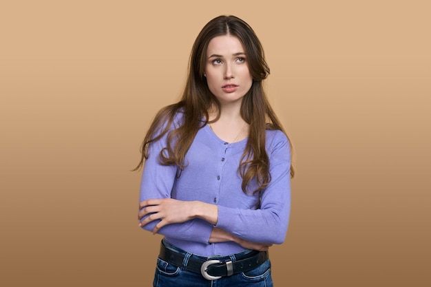 Pensive young woman dreamily looking aside standing with arms folded isolated on beige background