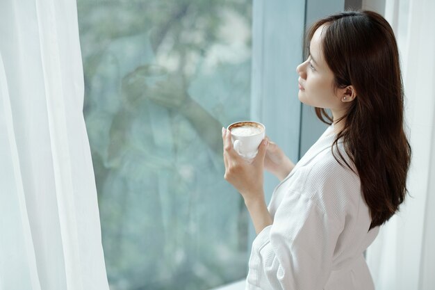 Photo pensive young woman drinking cup of morning coffee and looking outside through apartment window