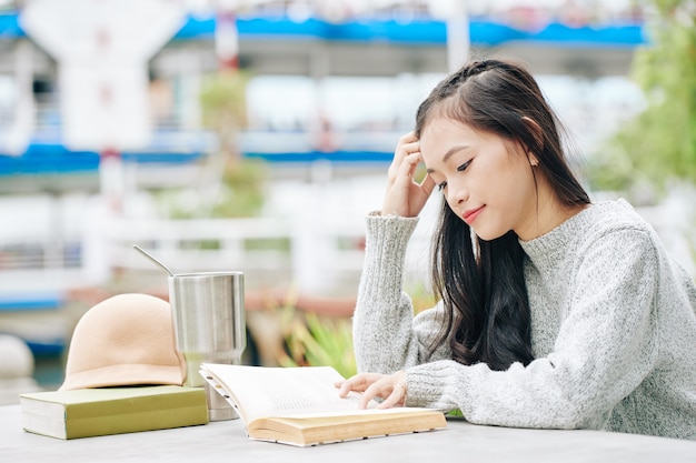 再利用可能なカップから飲んで、屋外で面白い本を読んで物思いにふける若い女性