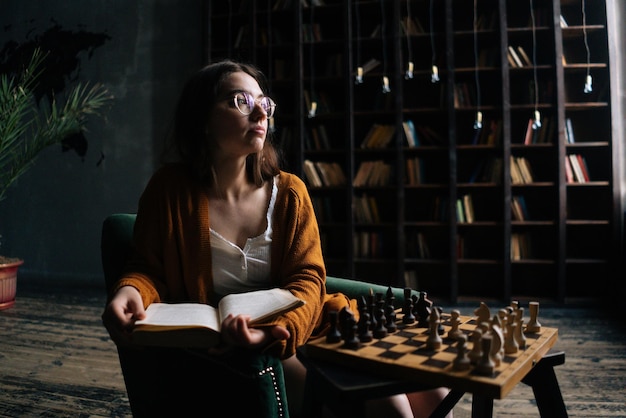 Giovane donna pensierosa in occhiali eleganti che tiene libro seduto in poltrona nella stanza buia della biblioteca pensando di distogliere lo sguardo sullo sfondo della libreria