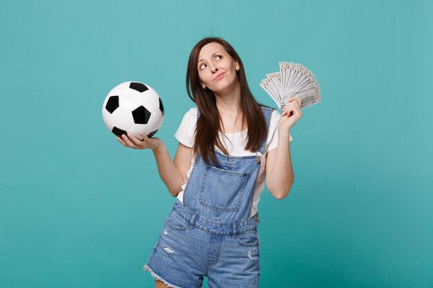 Pensive young woman football fan support favorite team with soccer ball, fan of money in dollar banknotes, cash money, looking up isolated on blue turquoise background. Sport family leisure concept.