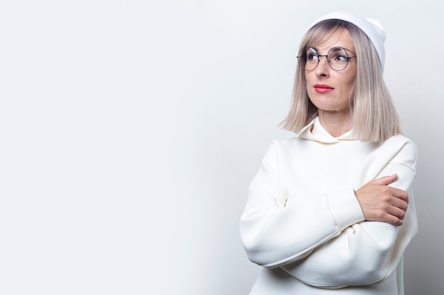 Pensive young woman in glasses with crossed arms on a light background.