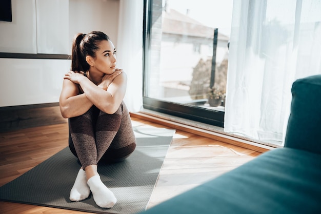 Pensive young woman is sitting on the yoga mat and preparing for training at home.