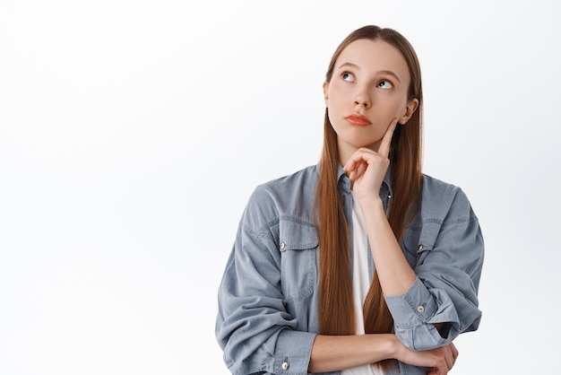 Pensive young woman with long hair look aside at upper left corner and thinking making choice pondering touching face thoughtful white background