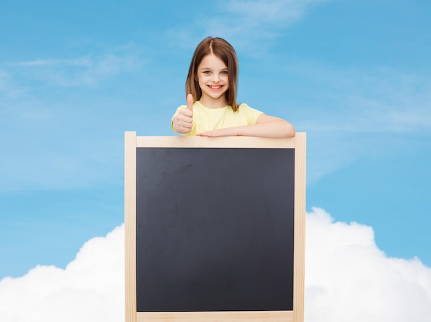 people, advertisement and education concept - happy little girl with blank blackboard showing thumbs up