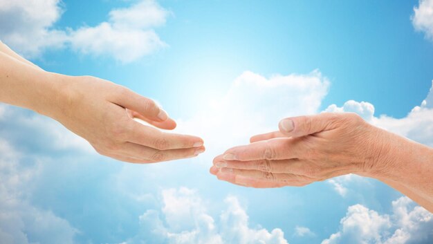 people, age, family, care and support concept - close up of senior woman and young woman reaching hands out to each other over blue sky and clouds background