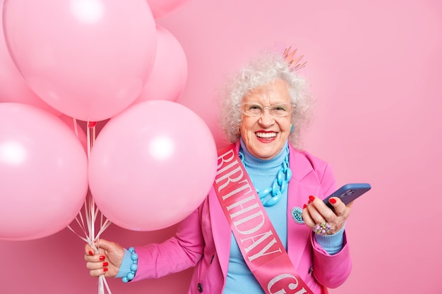 People age festive holiday concept. Positive good looking European female pensioner chuckles positively holds modern smartphone gets messages of congratultaion holds bunch of inflated balloons