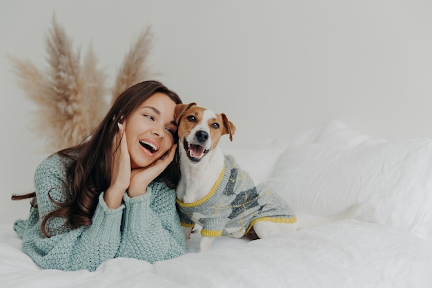 People and animals concept joyful pretty young woman express\
love to dog spends leisure time with pet lie together on bed looks\
tenderly at animal feels not bored with true loyal friend