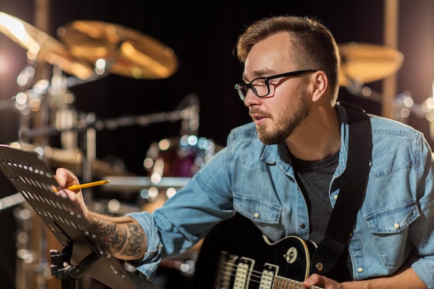 people, art, composition and entertainment concept - man with guitar writing lyrics or notes to music book at studio