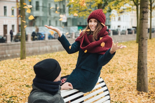 People autumn and relaxation concept Beautiful brunette female throws leaves looks joyfully at boyfriend spends weekends together in park dates Lovely couple in love enjoy warm autumn day