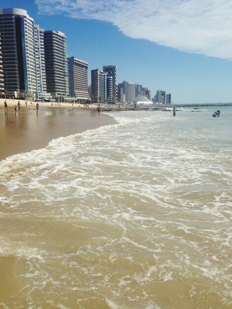 Photo people on beach by modern buildings against sky during sunny day