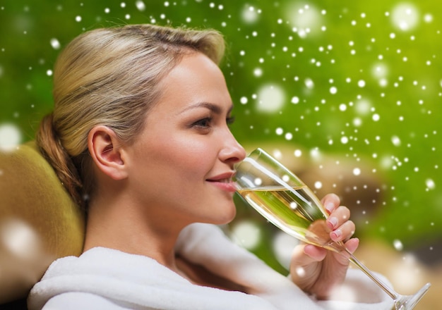 people, beauty, lifestyle, holidays and relaxation concept - beautiful young woman in white bath robe lying on chaise-longue and drinking champagne at spa with snow effect