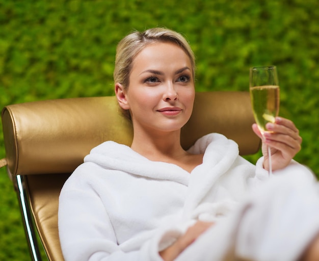 people, beauty, lifestyle, holidays and relaxation concept - beautiful young woman in white bath robe lying on chaise-longue and drinking champagne at spa