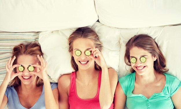 写真 人々、美しさ、自然化粧品、パジャマ パーティー コンセプト - 幸せな友人やキュウリ マスクを持つ 10 代の少女が楽しんで、家でベッドに横になっています。