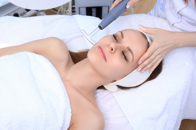 People, beauty, spa, cosmetology and technology concept - close up of beautiful young woman lying with closed eyes having face massage by massager in spa.