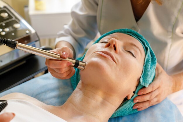 Photo people, beauty, spa, cosmetology and technology concept - cosmetologist makes the procedure microcurrent therapy of the facial skin of a beautiful, young woman in a beauty salon