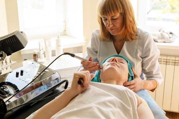 Photo people, beauty, spa, cosmetology and technology concept - cosmetologist makes the procedure microcurrent therapy of the facial skin of a beautiful, young woman in a beauty salon