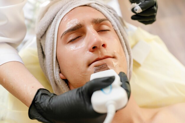 People, beauty, spa, cosmetology and technology concept - The cosmetologist makes the procedure ultrasonic face peeling of the facial skin of a beautiful, young man in a beauty salon