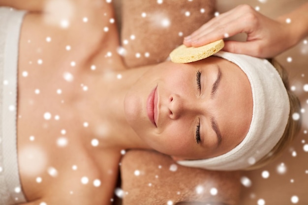 people, beauty, spa, healthy lifestyle and relaxation concept - close up of beautiful young woman lying with closed eyes and having face massage with sponge in spa with snow effect