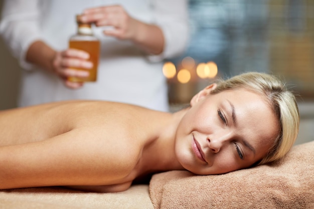 people, beauty, spa, healthy lifestyle and relaxation concept - close up of beautiful young woman lying with closed eyes on massage table and therapist holding oil bottle in spa