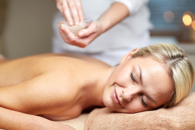 people, beauty, spa, massage and relaxation concept - close up of beautiful young woman lying with closed eyes and therapist holding salt bowl in spa