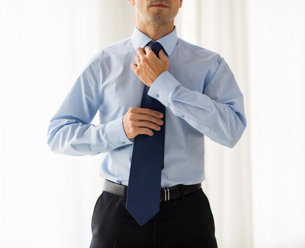 люди, бизнес, мода и концепция одежды - крупный план человека в рубашке, наряжающегося и поправляющего галстук на шее дома