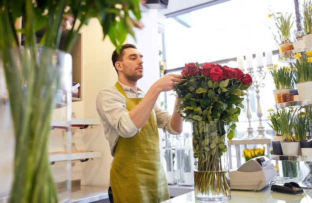 people, business, sale and floristry concept - happy smiling florist man with red roses at flower shop