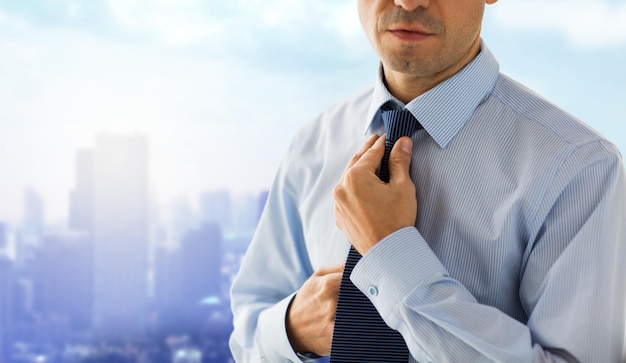 people, business, work and clothing concept - close up of man in shirt dressing up and adjusting tie on neck over city background
