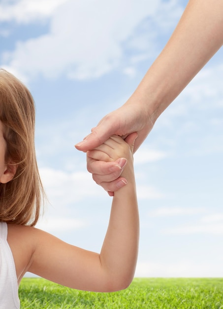 people, charity, family and adoption concept - close up of woman and little girl holding hands over blue sky and grass background