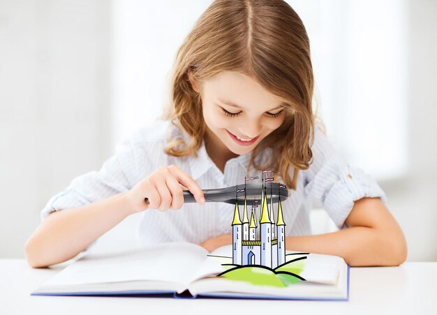 people, childhood, literature and imagination concept - little girl with book looking to fairy castle through magnifier at home