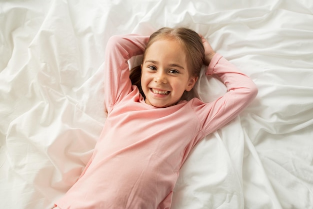 People, children and bedtime concept - happy little girl lying in bed at home