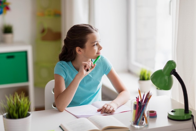 people, children, education and learning concept - happy girl with book and notebook looking through window at home