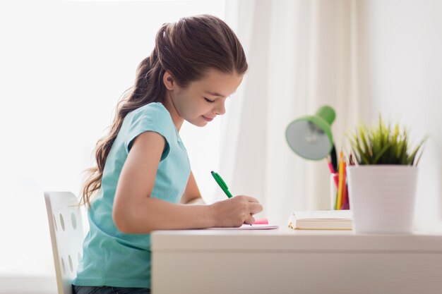 people, children, education and learning concept - happy girl with book writing to notebook at home