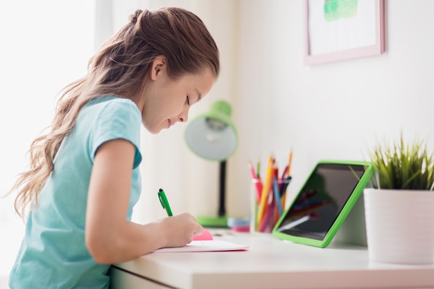 people, children, education and learning concept - happy girl with tablet pc computer writing to notebook at home